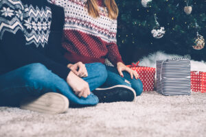 The Carpet Cleaners - Crop faceless couple in sweaters sitting on carpet near decorated fir tree with presents below.